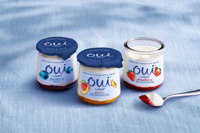 Three glass pots of Oui by Yoplait yogurt in Blueberry, Peach and Strawberry flavors sitting on a blue cloth. The Strawberry pot has the lid off and a spoon with the yogurt on it is sitting beside it.