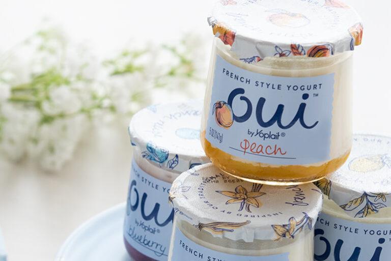 A jar of peach Oui by Yoplait yogurt stacked on other jars next to white flowers.