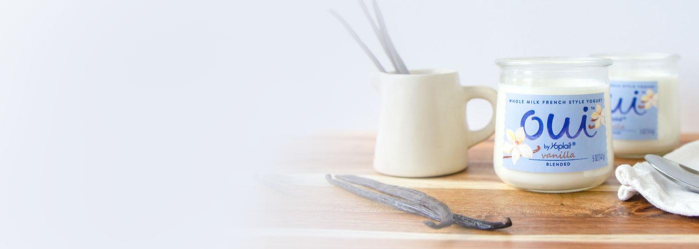 A jar of Vanilla Oui by Yoplait yogurt on a wood surface next to raw vanilla bean pods.