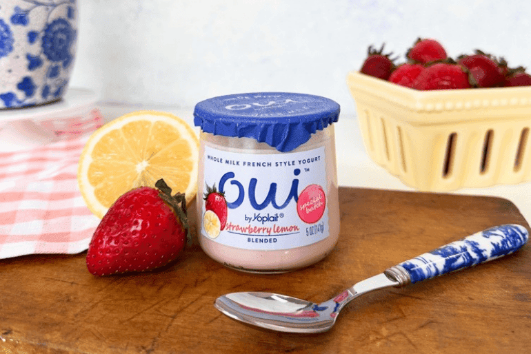 Close-up shot of a jar of OUI by Yoplait Strawberry Lemon blended yogurt on a rustic wooden cutting board. A ripe strawberry and a lemon wedge are positioned beside the yogurt jar. A spoon with a blue and white patterned handle is also on the board. A partially visible pink and white checkered napkin and a pale yellow basket of strawberries are in the background.